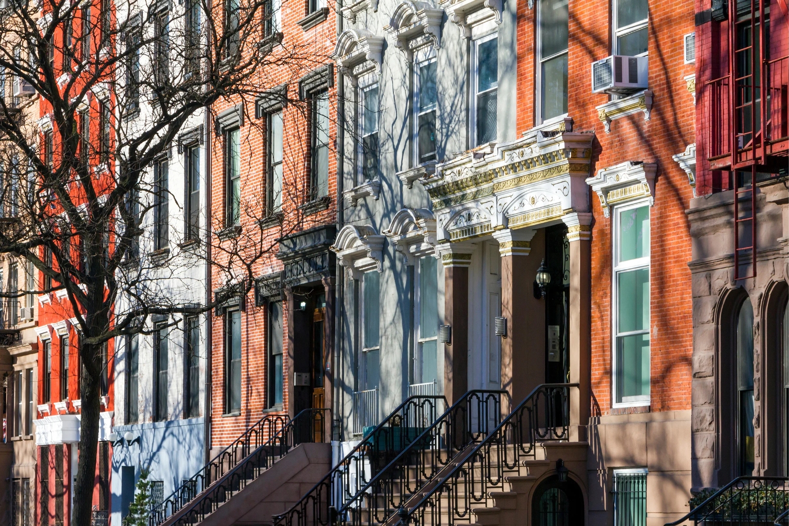 Sunlit row of historic brick and brownstone residential buildings with iron-railed stoops